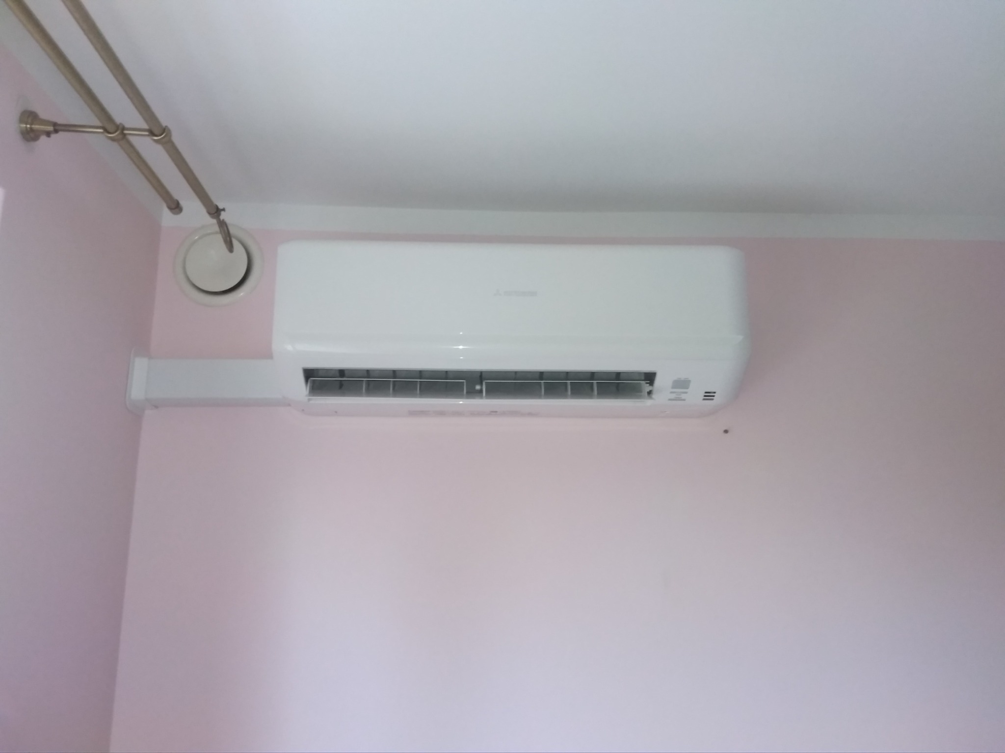 Biały klimatyzator Mitsubishi zamontowany na jasnoróżowej ścianie w pomieszczeniu z widocznym fragmentem instalacji wentylacyjnej pod sufitem.