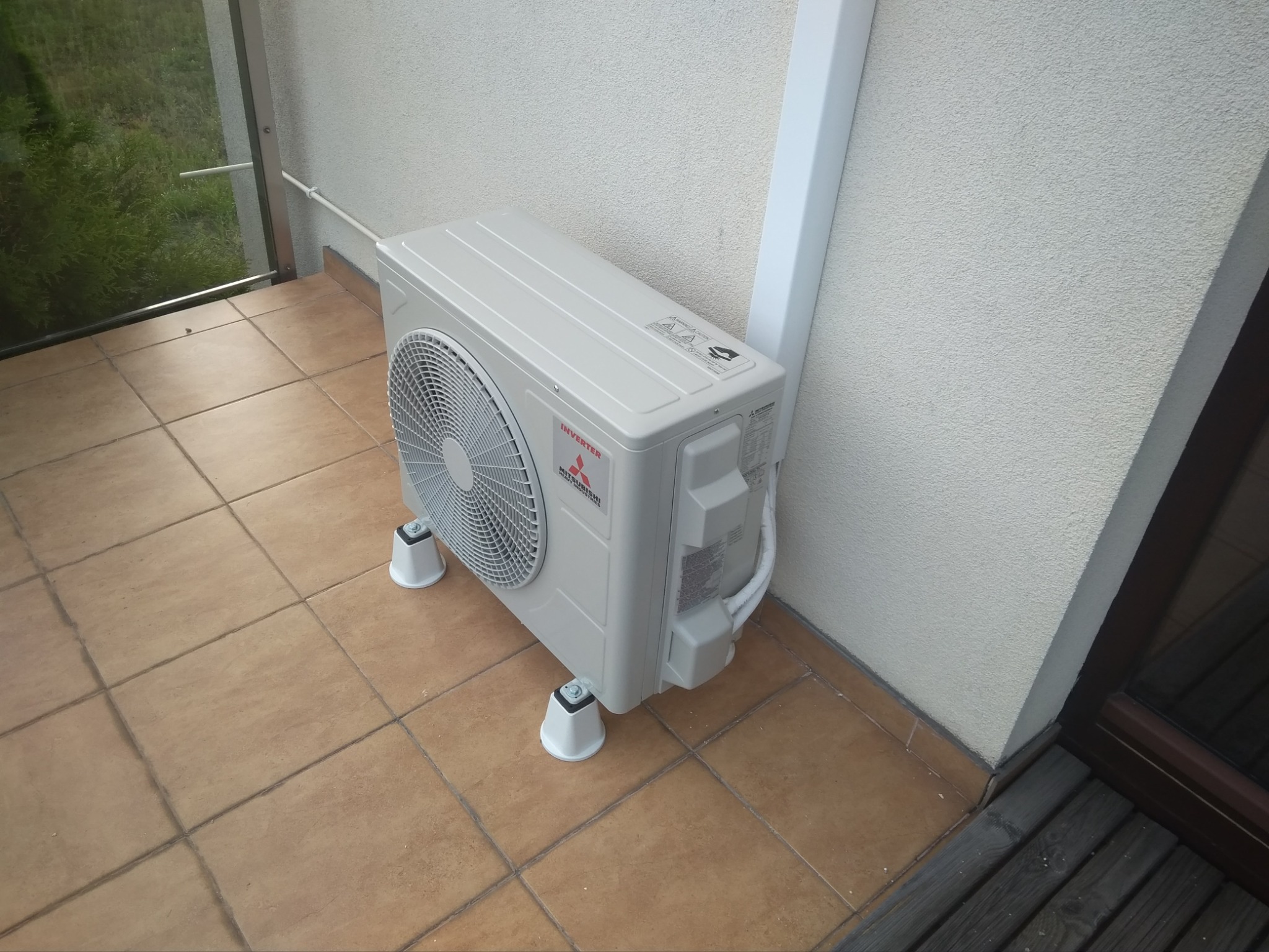 Zewnętrzna jednostka klimatyzacji Mitsubishi Inverter zamontowana na brązowych płytkach na balkonie, widoczne białe nóżki podtrzymujące urządzenie i biała rura odprowadzająca przewody.