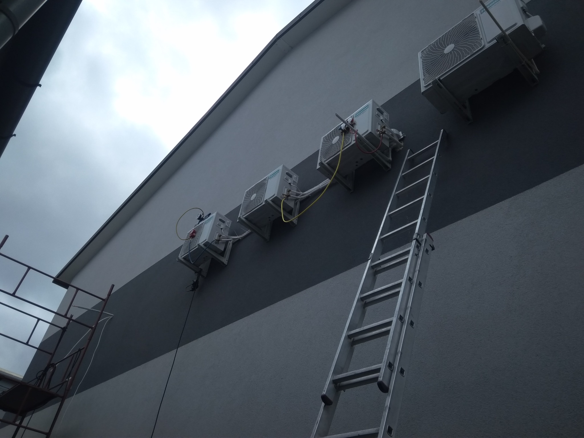 Zewnętrzne jednostki klimatyzacyjne zamontowane rzędem na dwukolorowej elewacji budynku, z aluminiową drabiną opartą o ścianę i fragmentem rusztowania w kadrze.