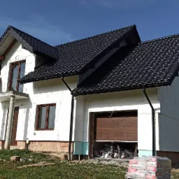 Dom jednorodzinny w trakcie ocieplania, widoczne białe płyty izolacyjne na elewacji, czarny dach z blachodachówki, częściowo otwarta brama garażowa, worki z zaprawą na pierwszym planie.