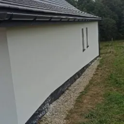 Nowoczesny dom jednorodzinny z jasną elewacją, ciemnym dachem i wąskimi oknami, otoczony zielenią i kamiennym drenażem.