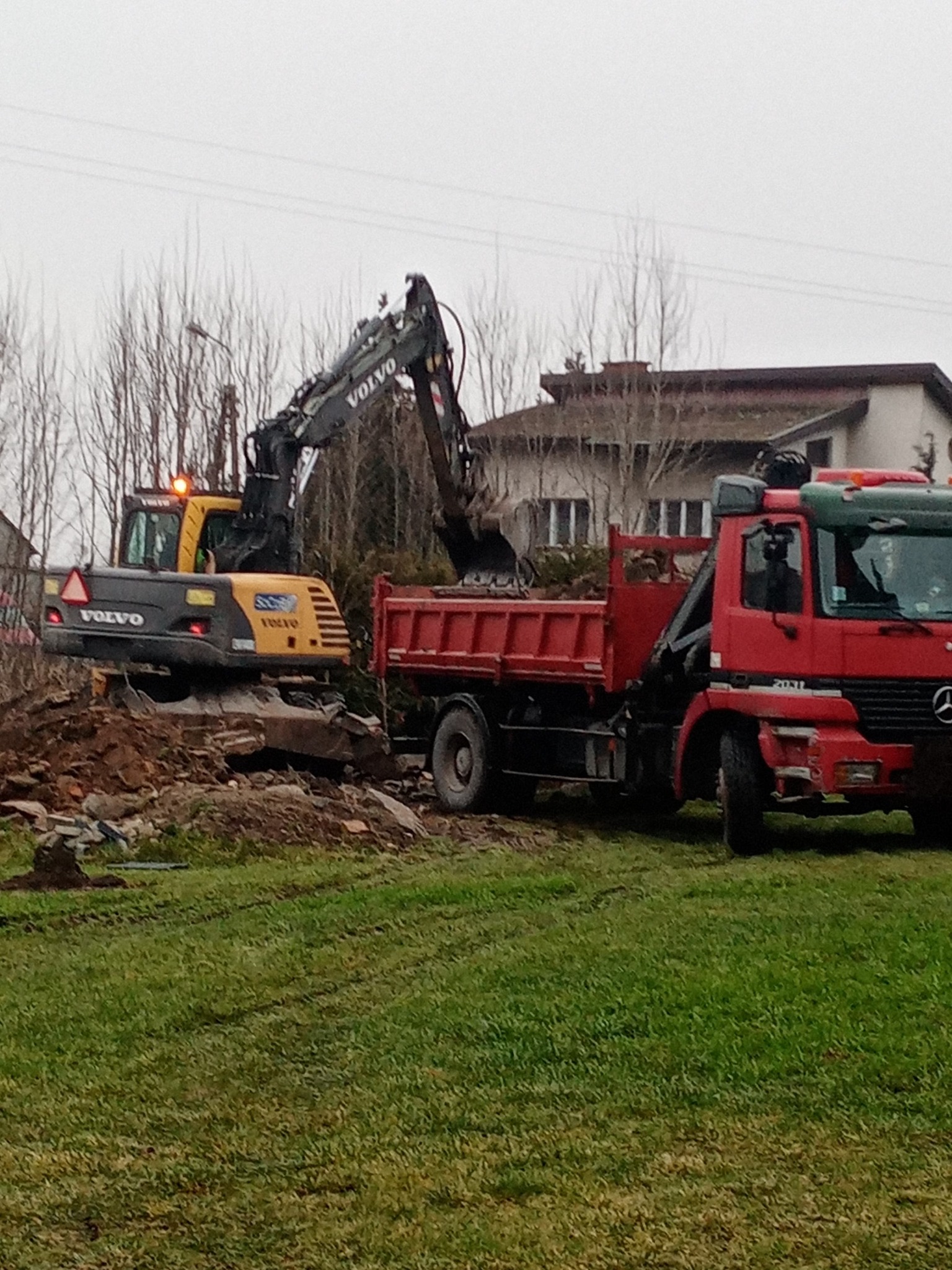 Żółto-szara koparka Volvo ładuje gruz do czerwonej ciężarówki na tle pochmurnego nieba i zabudowań.
