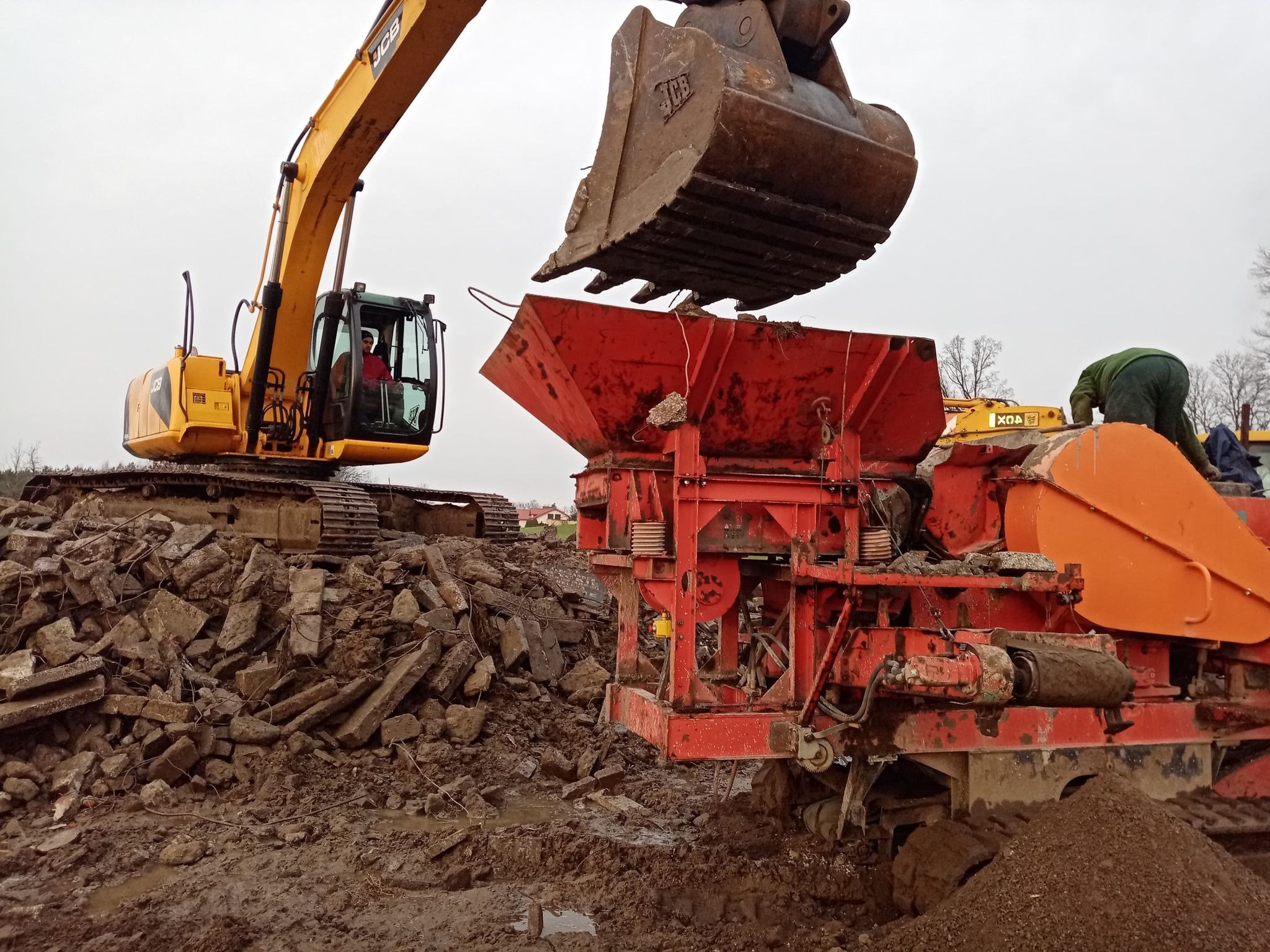 Żółta koparka JCB z operatorem w kabinie, przesypuje gruz do czerwonej kruszarki na błotnistym terenie rozbiórki, w tle widoczne zabudowania.