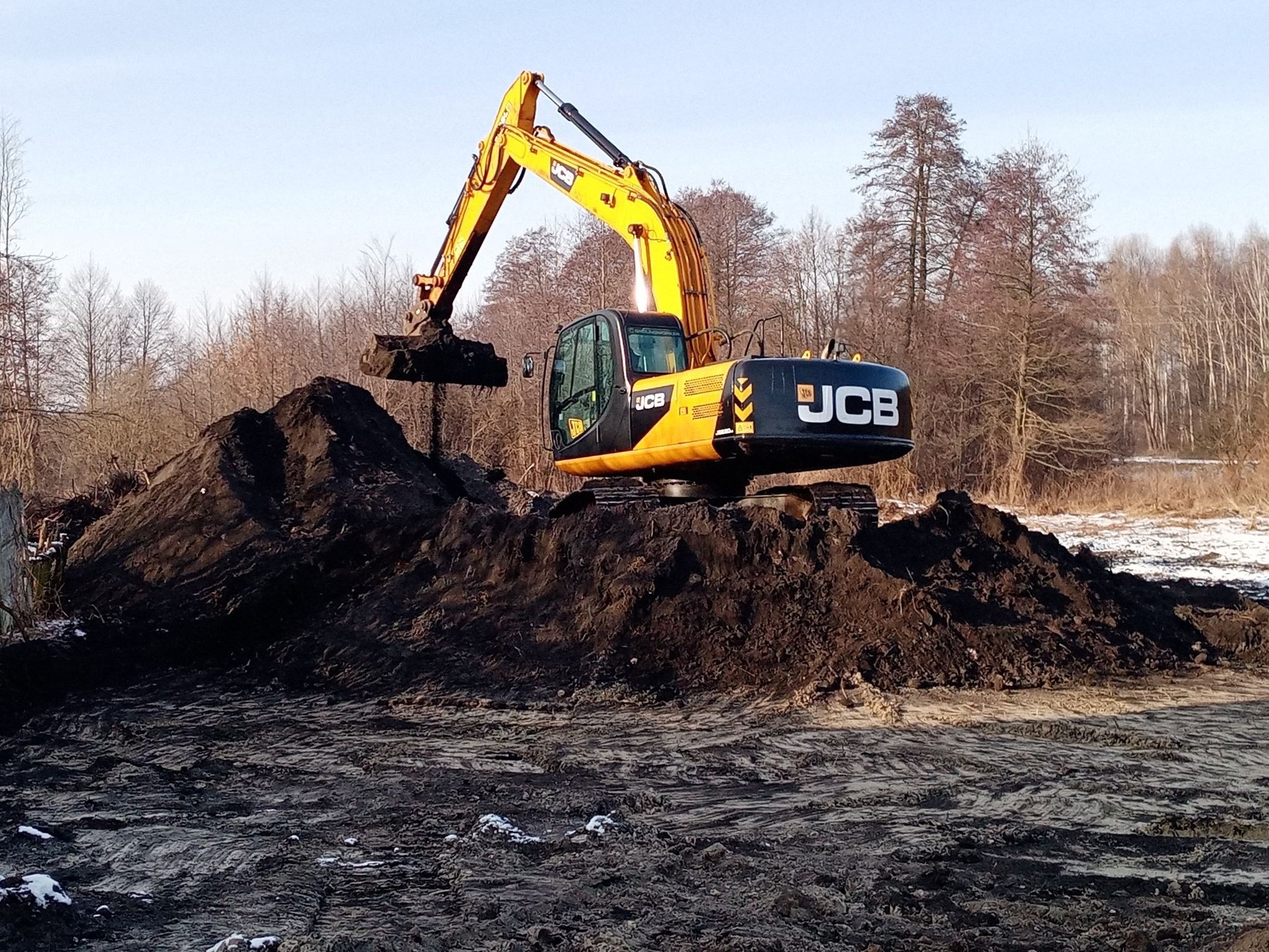 Żółto-czarna koparka JCB nabiera ziemię z pryzmy na tle zimowego krajobrazu z drzewami i resztkami śniegu.