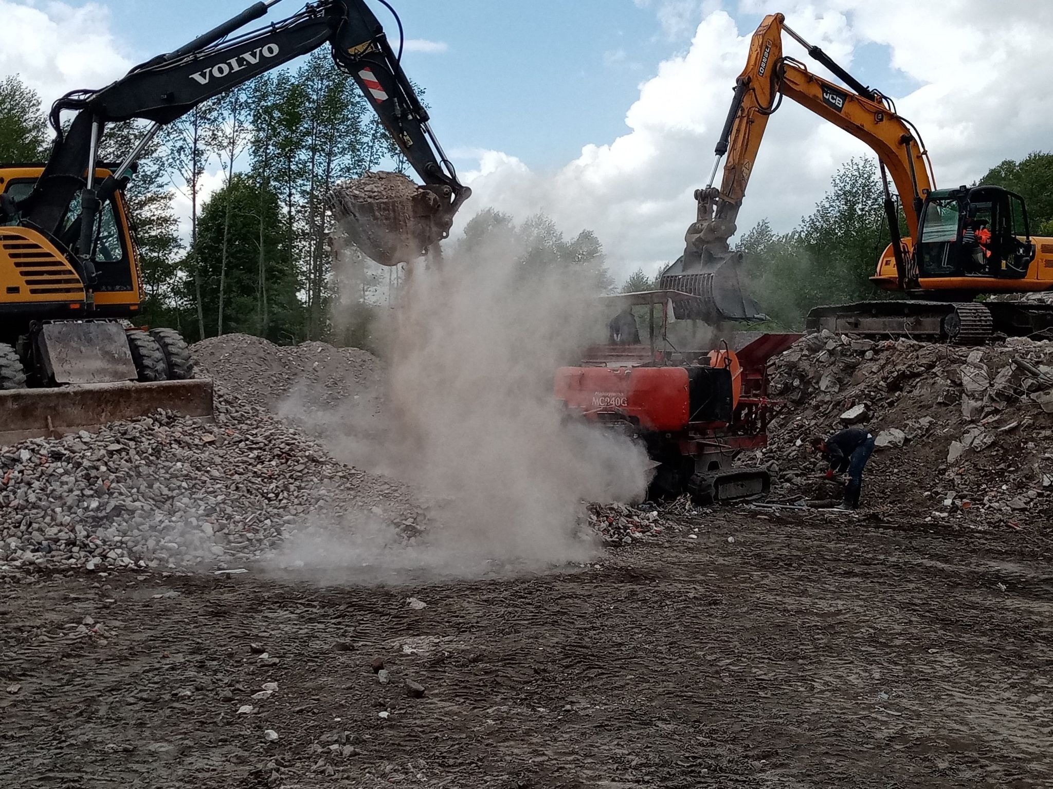 Dwa żółte i czarne koparki Volvo i JCB pracują przy kruszarce do gruzu na placu budowy, unosząc tumany kurzu, w tle widoczny pracownik w ciemnym ubraniu.