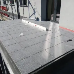 Szare płytki elewacyjne ułożone na balkonie budynku, widoczne poziomice i krzyżyki dystansowe, rusztowanie w tle.