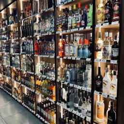 LK Polska Sp. z o. o. - Półki w sklepie z alkoholem, wypełnione różnorodnymi butelkami wódek, likierów, rumów i innych trunków, podświetlone ciepłym światłem, z widocznymi cenami na etykietach.