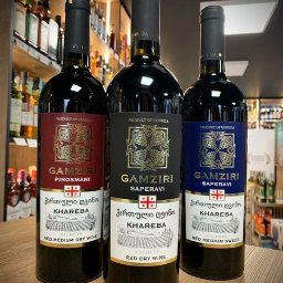 LK Polska Sp. z o. o. - Trzy butelki gruzińskiego wina Gamziri Khareba: czerwone półwytrawne Pirośmani, czerwone wytrawne Saperavi i czerwone słodkie Saperavi, ustawione na drewnianym blacie na tle regałów z alkoholem.