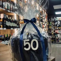 Czarny tort urodzinowy z liczbą 50, zapakowany w przezroczysty celofan i przewiązany niebieską wstążką, na drewnianym blacie, w tle półki z butelkami alkoholi.