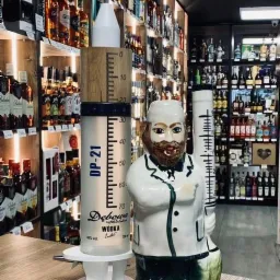 Dwie dekoracyjne butelki na alkohol: jedna w kształcie strzykawki z podziałką, marki Dębowa, druga w kształcie lekarza w białym fartuchu, na tle półek z różnymi alkoholami w sklepie.