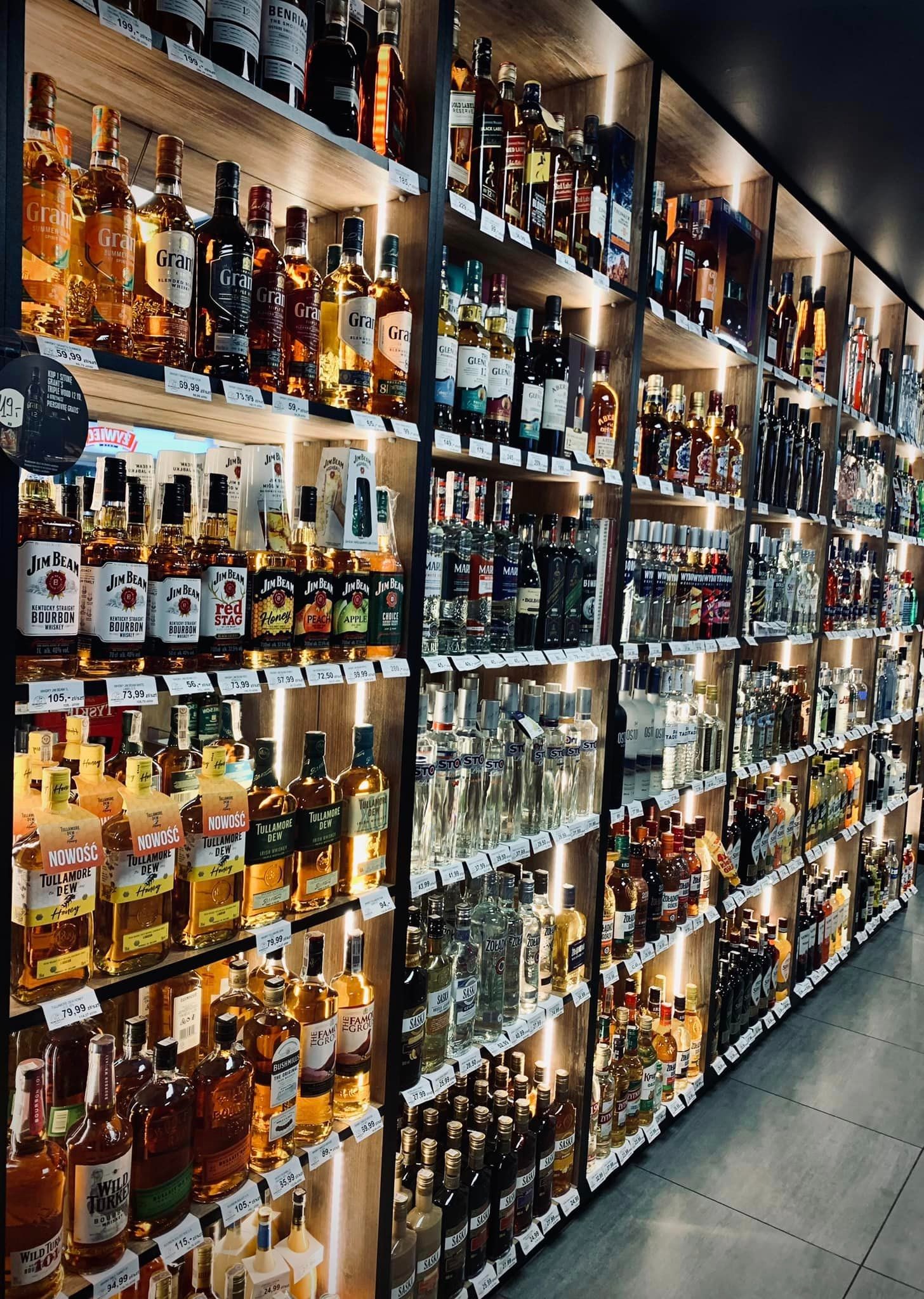 Półki w sklepie z szerokim wyborem butelek alkoholi, w tym whisky, bourbon, wódki i likiery, z cenami na półkach.