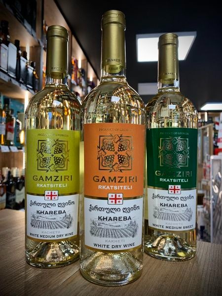 Trzy butelki gruzińskiego białego wina Gamziri Rkatsiteli o różnych etykietach (żółta, pomarańczowa, zielona) stojące obok siebie na drewnianym blacie, na tle półek sklepowych z innymi alkoholami.