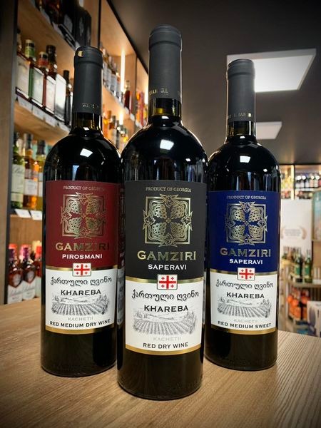 Trzy butelki gruzińskiego wina Gamziri Khareba: czerwone półwytrawne Pirośmani, czerwone wytrawne Saperavi i czerwone słodkie Saperavi, ustawione na drewnianym blacie na tle regałów z alkoholem.
