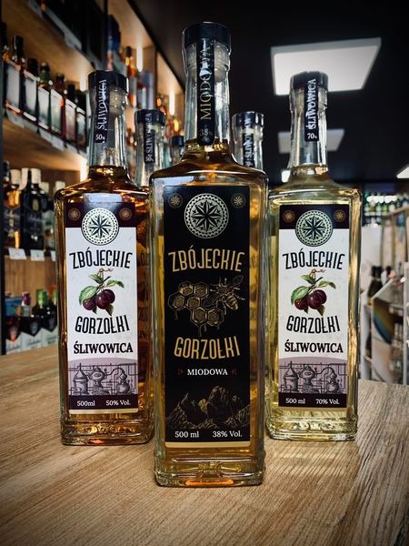 Trzy butelki regionalnego alkoholu Zbojeckie Gorzołki: śliwowica 50%, miodowa 38% i śliwowica 70%, ustawione na drewnianym blacie w sklepie z alkoholem.