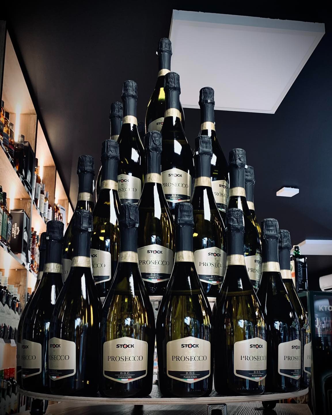 Piramida ułożona z butelek Prosecco marki Stock, z widocznymi etykietami i czarnymi kapslami, na tle półek z innymi alkoholami i czarnego sufitu z oświetleniem.