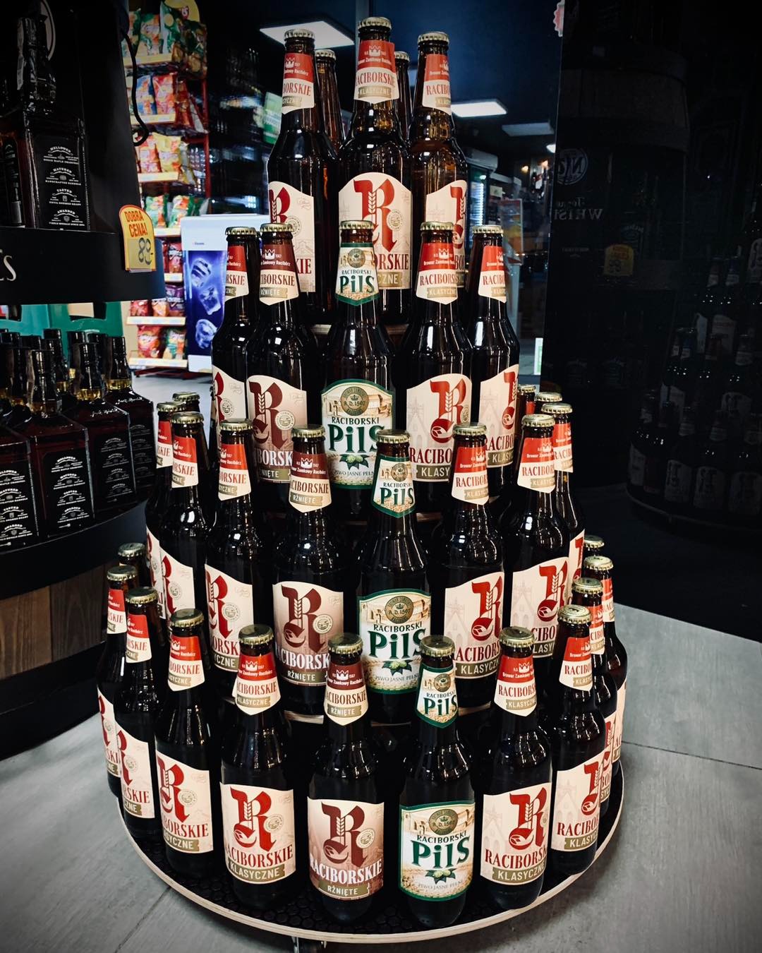 Piramida ułożona z butelek piwa Raciborskie Klasyczne i Pils na tle sklepowych półek z alkoholem.