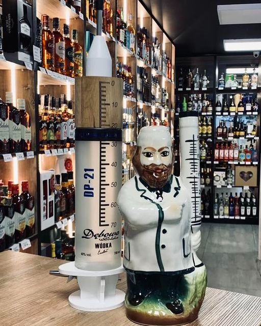 Dwie dekoracyjne butelki na alkohol: jedna w kształcie strzykawki z podziałką, marki Dębowa, druga w kształcie lekarza w białym fartuchu, na tle półek z różnymi alkoholami w sklepie.