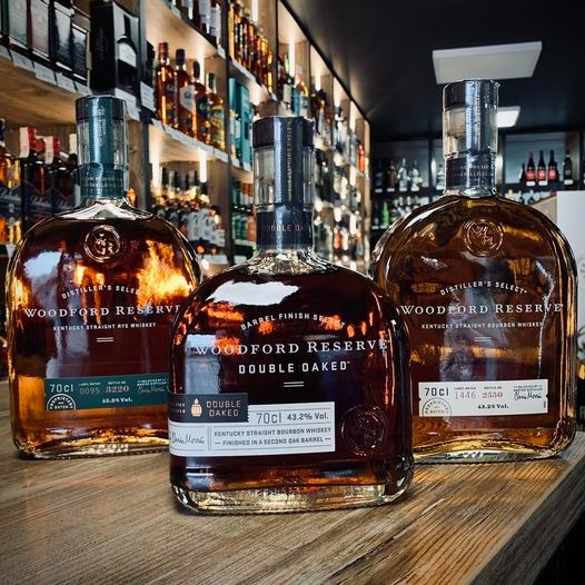 Trzy butelki Woodford Reserve, w tym Double Oaked, ustawione na drewnianym blacie na tle półek z alkoholem w sklepie.