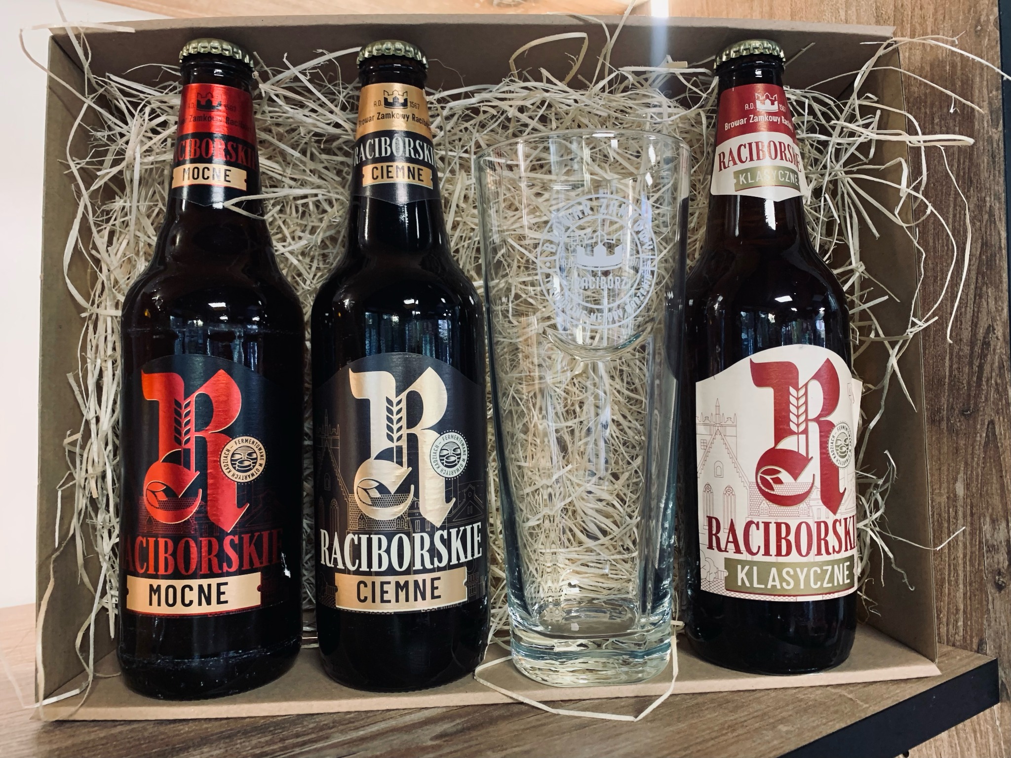 Trzy butelki piwa Raciborskie (Mocne, Ciemne, Klasyczne) oraz szklanka z logo browaru, ułożone w tekturowym pudełku wypełnionym siankiem dekoracyjnym.