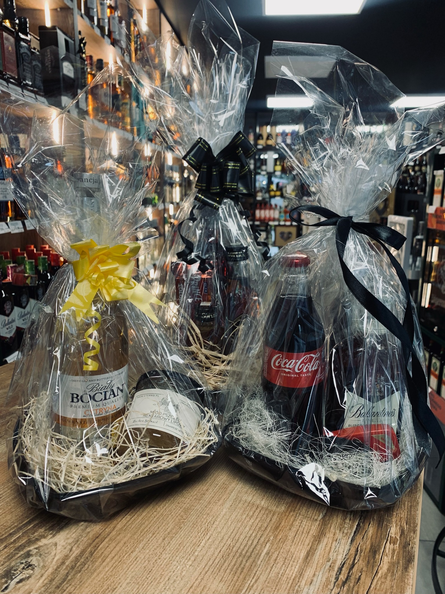 Dwa kosze prezentowe z alkoholami i napojami, zapakowane w celofan i ozdobione wstążkami, ustawione na drewnianym blacie na tle półek z butelkami w sklepie.