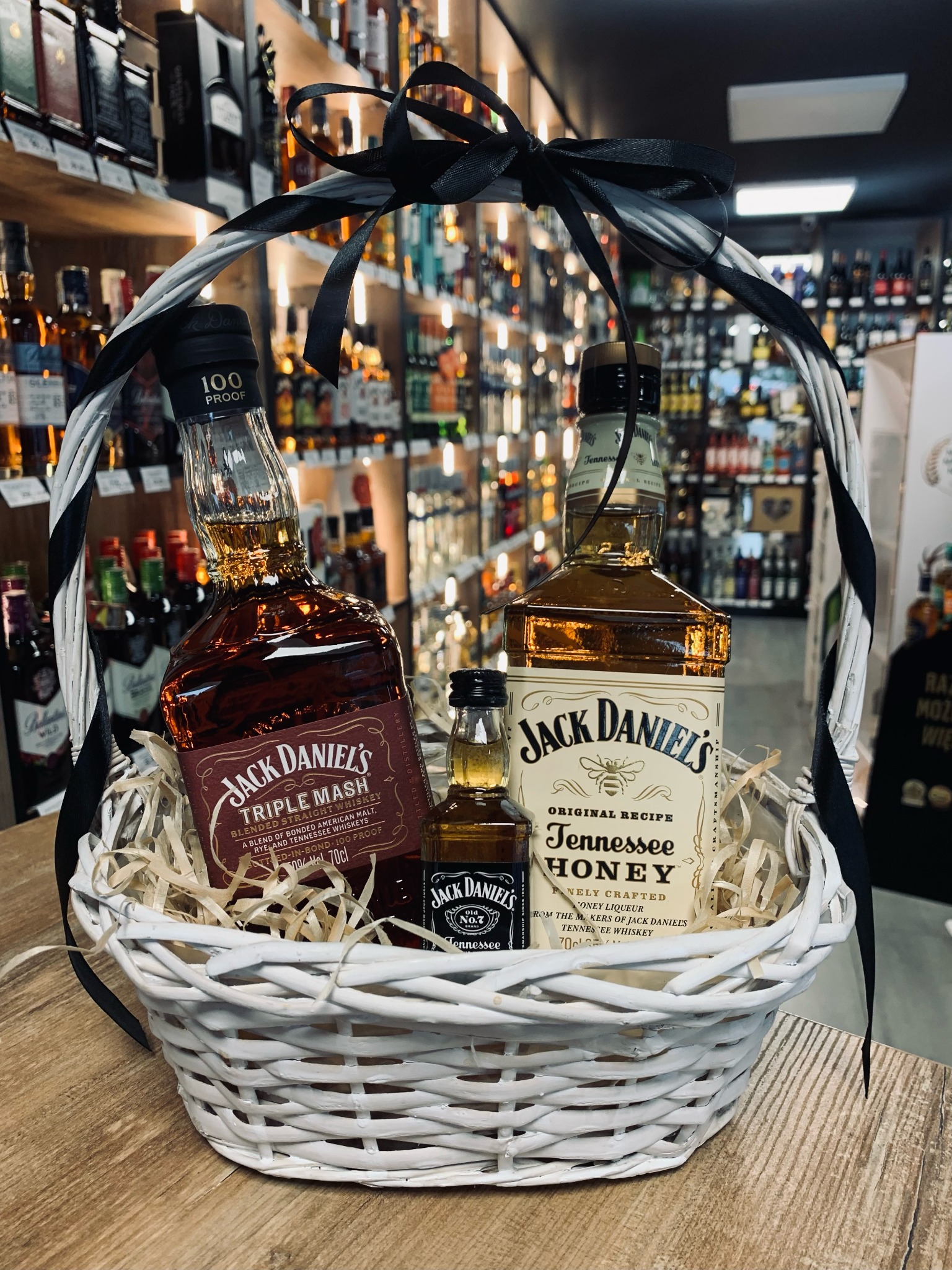 Biały wiklinowy kosz prezentowy wypełniony trzema butelkami Jack Daniel's (Triple Mash, Tennessee Honey i mała butelka No. 7), ozdobiony czarną wstążką, na tle półek sklepowych z alkoholem.