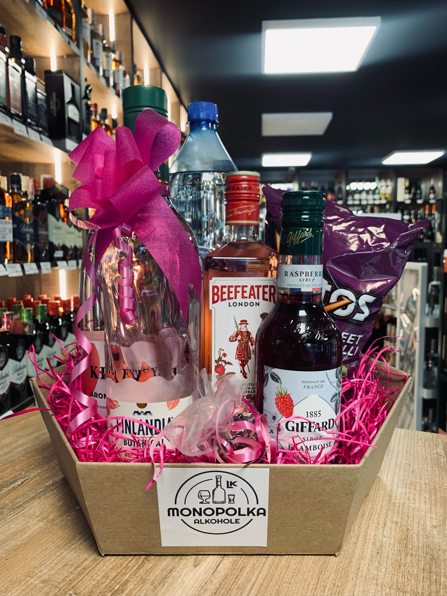Kosz prezentowy z różową wstążką, zawierający butelki alkoholu (Finlandia, Beefeater) oraz syrop malinowy Giffard, umieszczony na tle półek sklepowych z alkoholem.