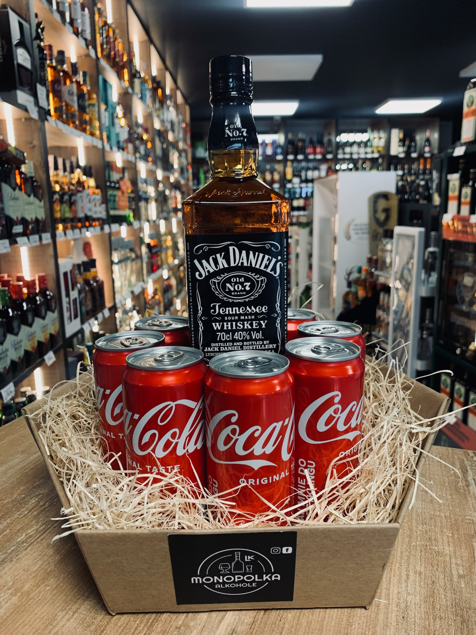 Kartonowy kosz prezentowy wypełniony wełną drzewną, zawierający butelkę Jack Daniel's i sześć puszek Coca-Coli, na tle półek sklepowych z alkoholem. Na koszu widoczne logo 'Monopolka Alkohole'.