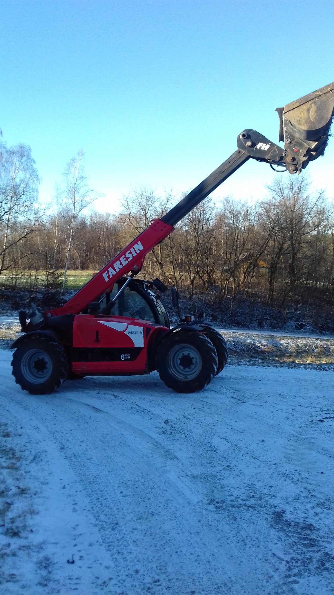 Czerwona ładowarka teleskopowa Faresin 632 z łyżką, stojąca na ośnieżonej drodze, z lasem w tle i błękitnym niebem.