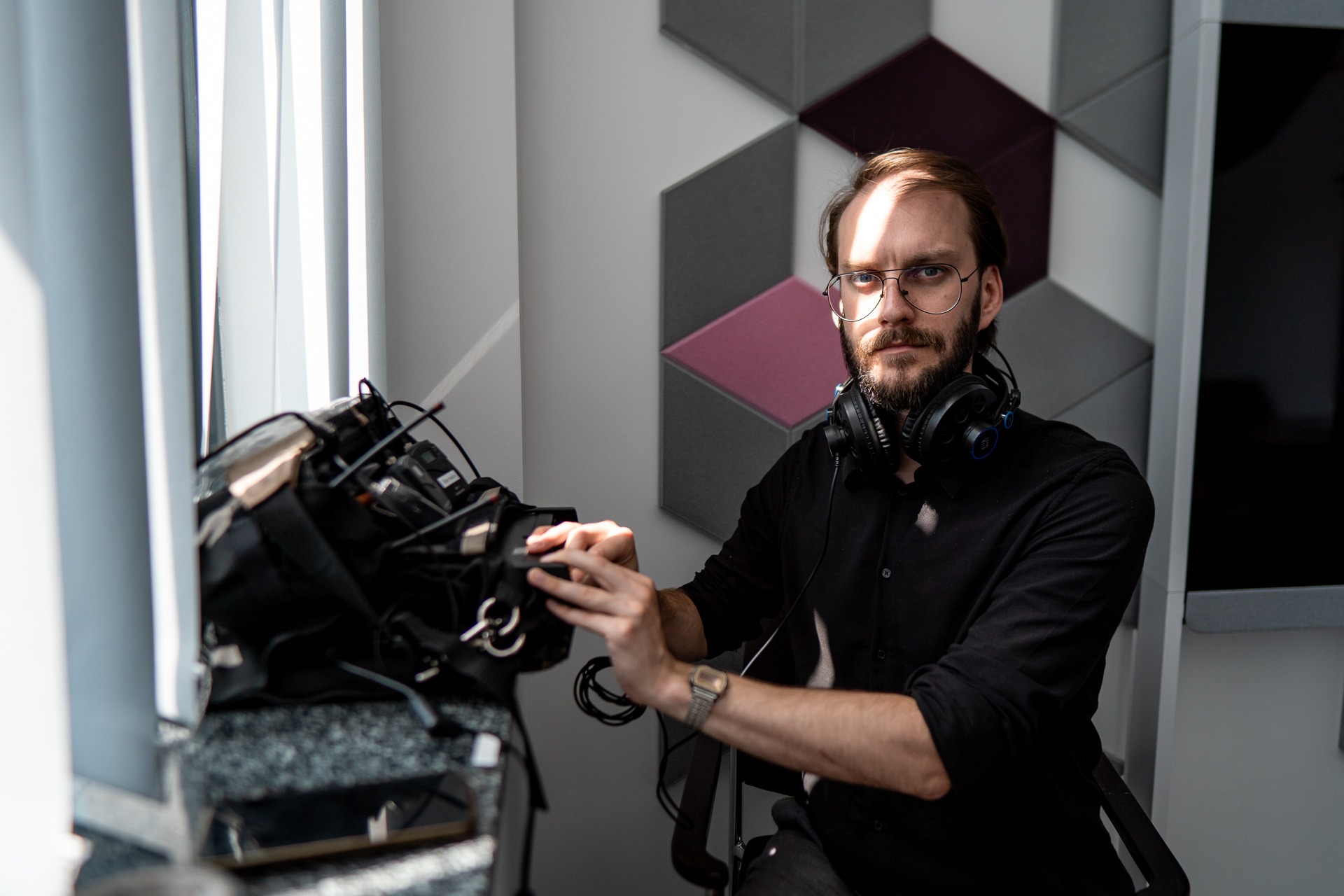 Mężczyzna w okularach i słuchawkach, siedzący obok torby z mikrofonami i rejestratorami dźwięku, w pomieszczeniu z panelami akustycznymi na ścianie.
