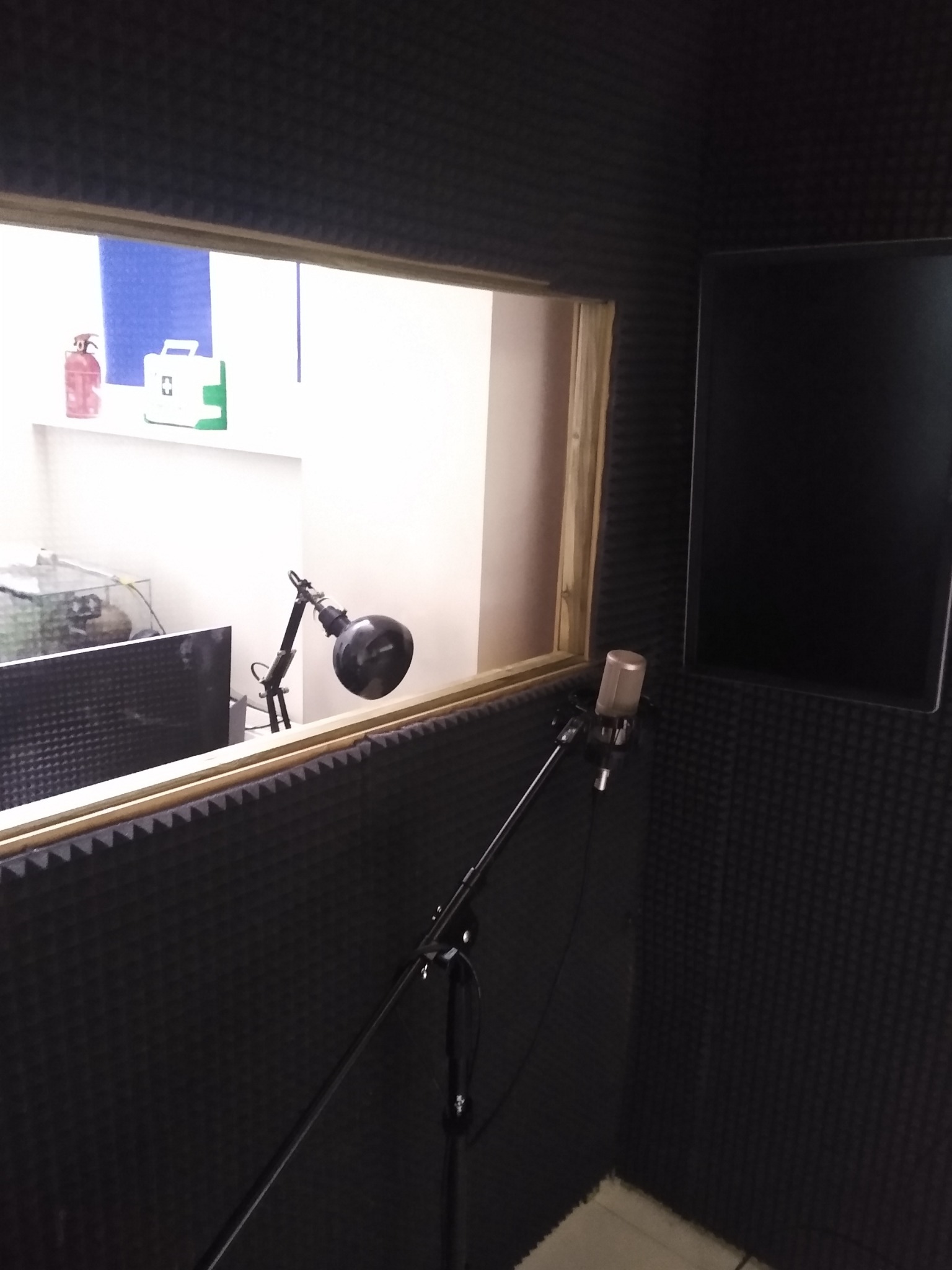 Wnętrze kabiny nagraniowej z mikrofonem na statywie, wygłuszone ściany, okno z widokiem na reżyserkę z lampką i monitorem, apteczka i gaśnica na parapecie.