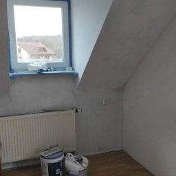 Sam-Bud - Pokój na poddaszu w trakcie remontu: ściany po wstępnym wyrównaniu, okno zabezpieczone taśmą malarską, widoczne dwa wiadra z farbą i grzejnik.