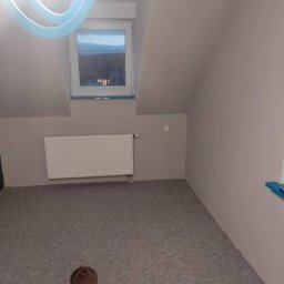 Sam-Bud - Poddasze po remoncie: świeżo pomalowane ściany w odcieniach szarości, nowy dywan, biały grzejnik pod oknem dachowym. Widoczny fragment nowoczesnej lampy sufitowej.