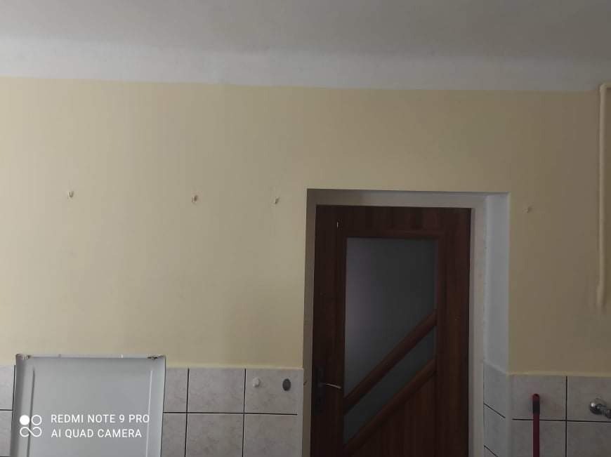 Pomieszczenie po malowaniu ścian na jasny, kremowy kolor, widoczne ślady po wcześniejszych mocowaniach na ścianie, brązowe drzwi z szybą i fragment białej lodówki.