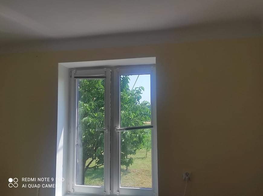 Pomalowane na beżowo ściany wewnętrzne z białymi ramami okiennymi, widok na drzewa za oknem, widoczne gniazdko elektryczne.