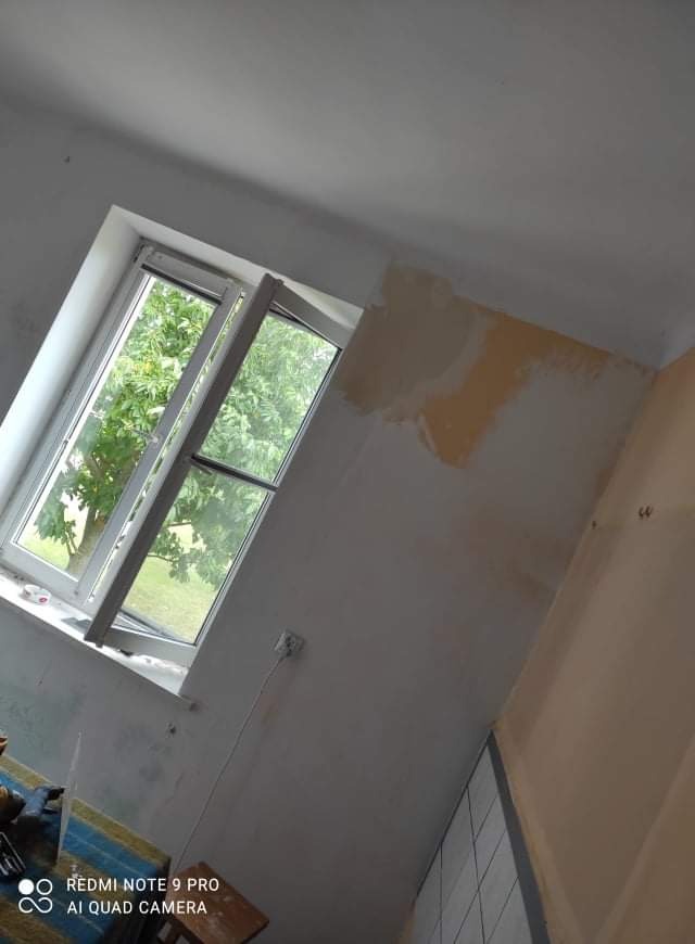 Ściana w trakcie prac przygotowawczych do malowania, widoczne fragmenty starej farby i nałożonej warstwy wyrównującej, otwarte okno z widokiem na drzewa.