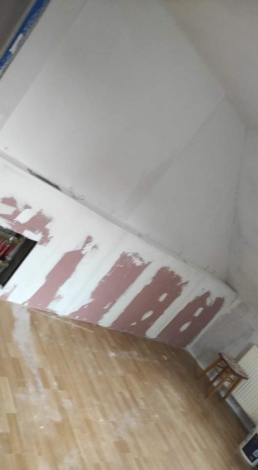 Skośna ściana poddasza w trakcie prac wykończeniowych, z widocznymi pasami nałożonej warstwy wyrównującej, przygotowana do dalszej obróbki, z fragmentem drewnianej podłogi w dolnej części kadru.