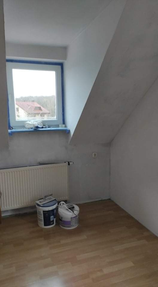 Pokój na poddaszu w trakcie remontu: ściany po wstępnym wyrównaniu, okno zabezpieczone taśmą malarską, widoczne dwa wiadra z farbą i grzejnik.