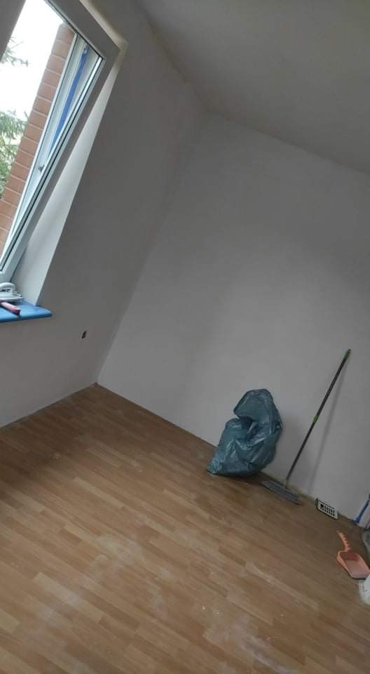 Pokój po świeżym malowaniu, z widoczną torbą na śmieci, miotłą i drobnymi zabrudzeniami na podłodze z paneli, okno dachowe po lewej stronie.