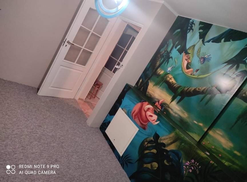 Pokój dziecięcy z motywem Króla Lwa na ścianie, widoczny fragment białych drzwi z przeszkleniami, szara wykładzina podłogowa i okrągła lampa sufitowa.