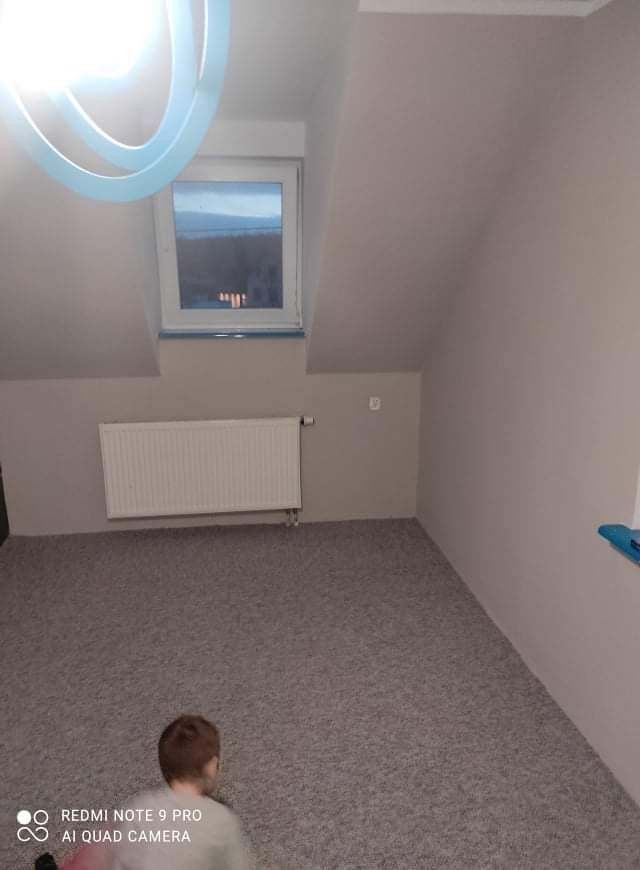 Poddasze po remoncie: świeżo pomalowane ściany w odcieniach szarości, nowy dywan, biały grzejnik pod oknem dachowym. Widoczny fragment nowoczesnej lampy sufitowej.