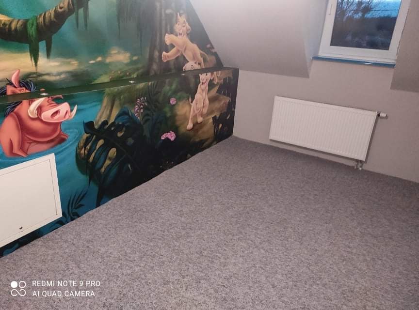 Pokój dziecięcy z szarą wykładziną, ściana z motywem Króla Lwa, okno dachowe i biały grzejnik.