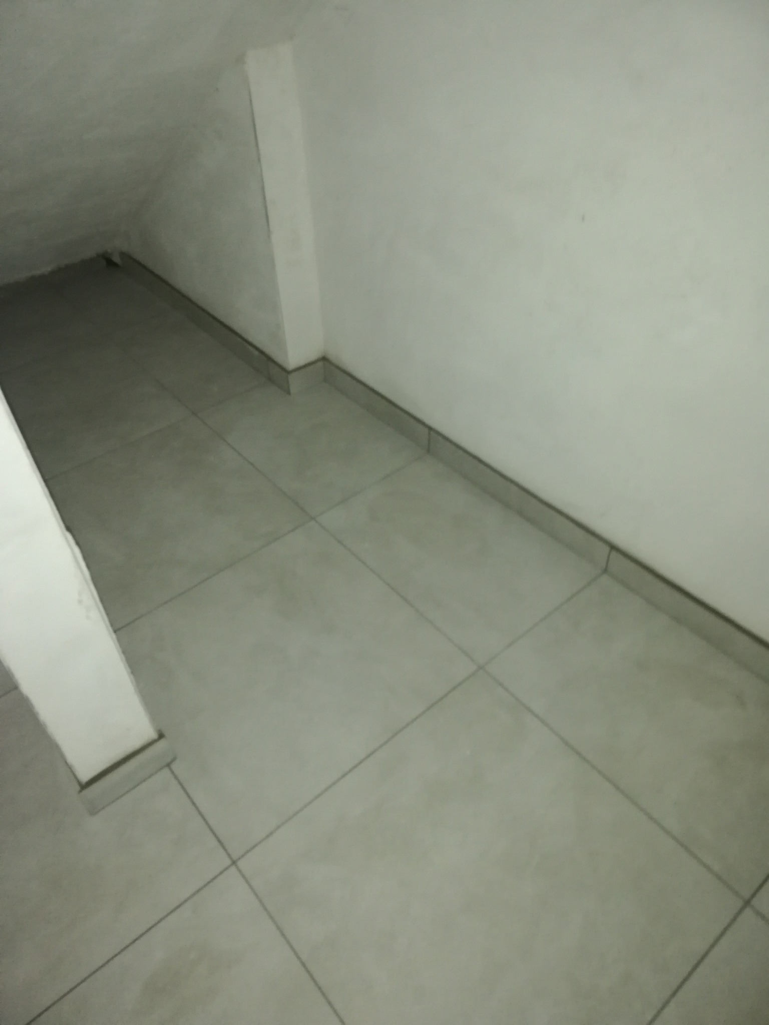 Szare, kwadratowe płytki podłogowe w pomieszczeniu ze skosem sufitowym i listwami przypodłogowymi, widoczne słabe oświetlenie