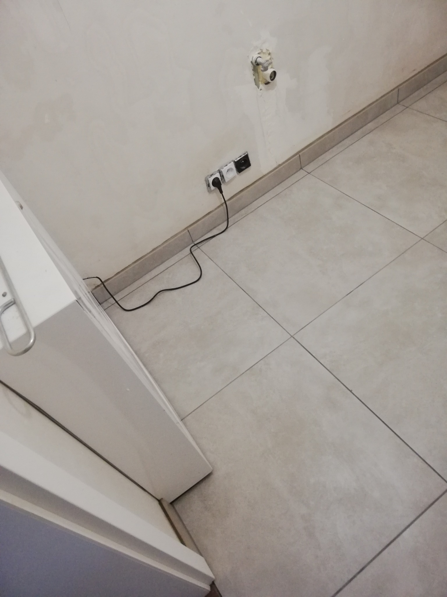 Jasne, kwadratowe płytki podłogowe ułożone w łazience, widoczne gniazdka elektryczne na ścianie i fragment białej umywalki.