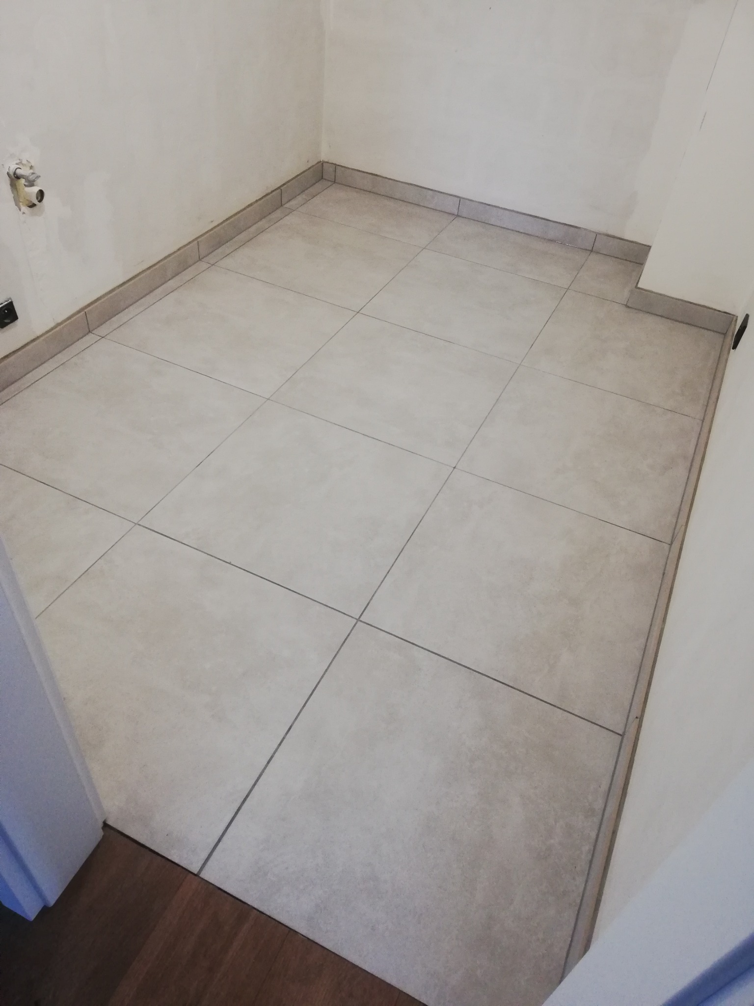 Wykończona podłoga w małym pomieszczeniu z jasnymi, kwadratowymi płytkami ceramicznymi ułożonymi równo, z widoczną listwą przypodłogową z płytek i niezamaskowaną puszką elektryczną na ścianie.