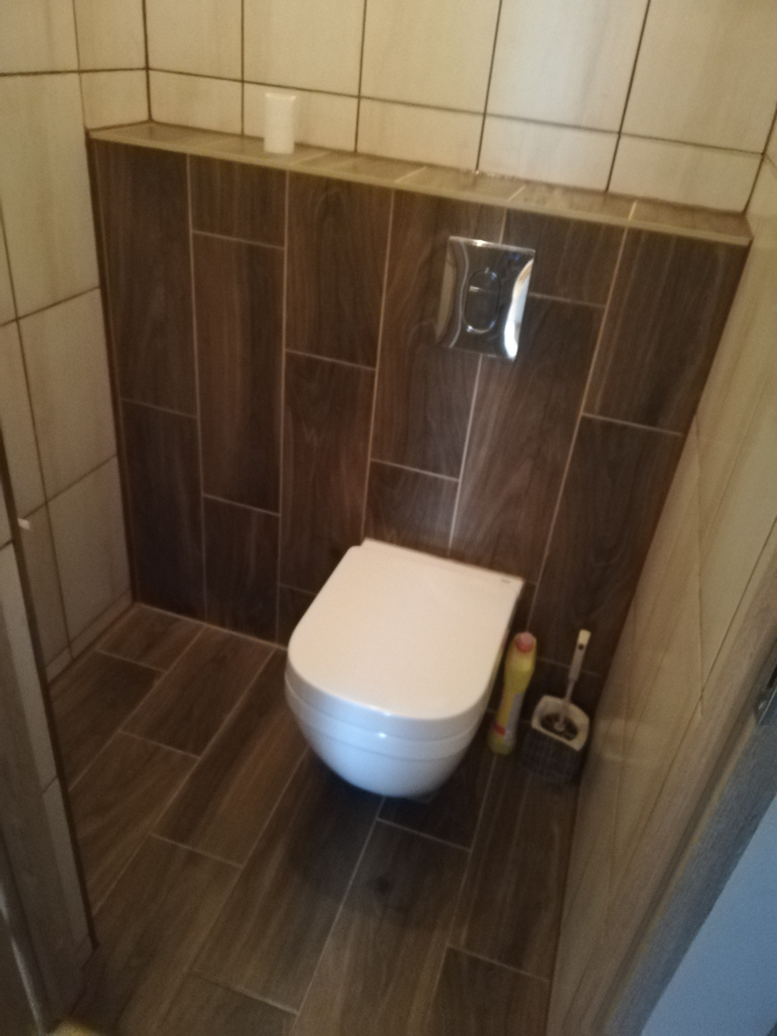 Wisząca toaleta z białą miską i deską, zamontowana na ścianie wyłożonej brązowymi płytkami imitującymi drewno, z chromowanym przyciskiem spłukującym. Podłoga również wyłożona płytkami w odcieniach...