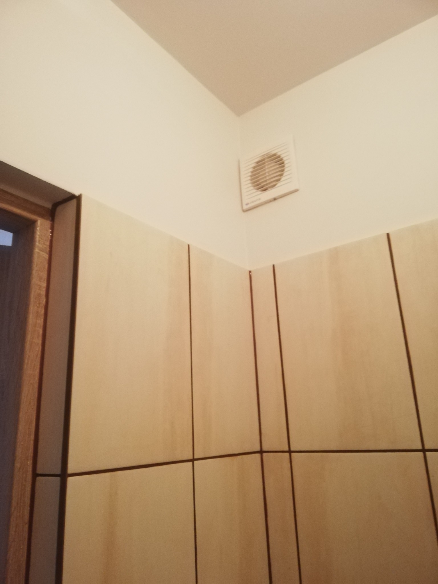 Ściana wyłożona jasnymi, kwadratowymi płytkami ceramicznymi z ciemnymi fugami, widoczny fragment drewnianej ościeżnicy drzwi i biały wentylator zamontowany na ścianie pod sufitem.