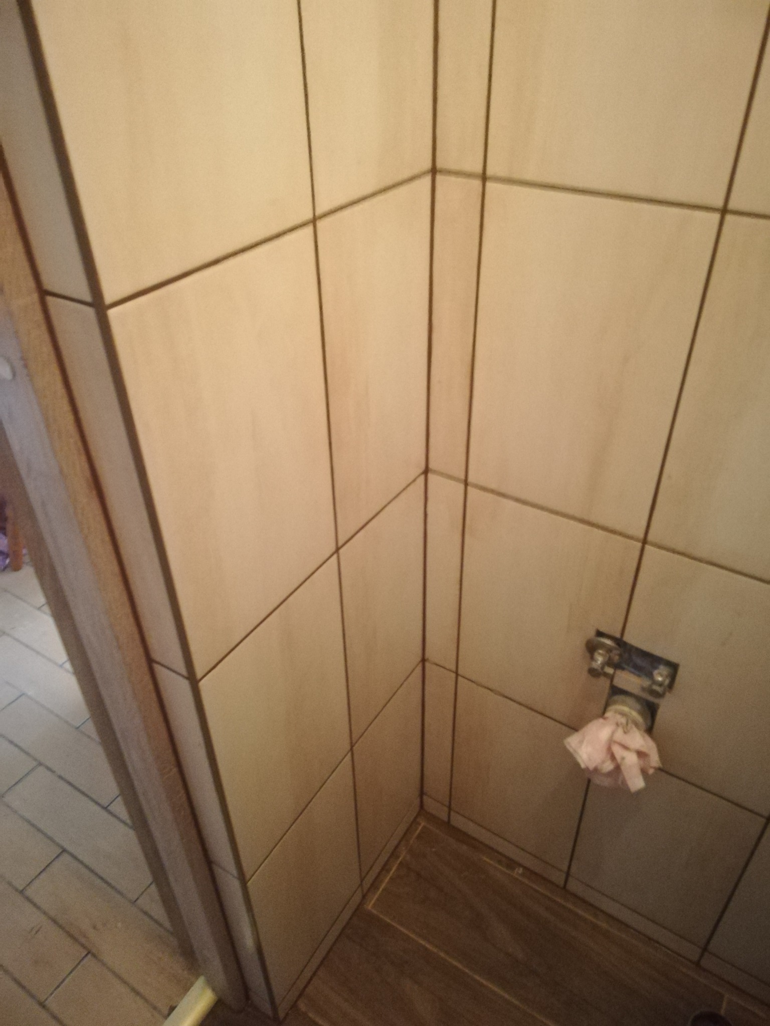 Narożnik wyłożony beżowymi płytkami ceramicznymi, widoczne mocowanie rury z zawiązaną szmatką, na podłodze płytki imitujące drewno.