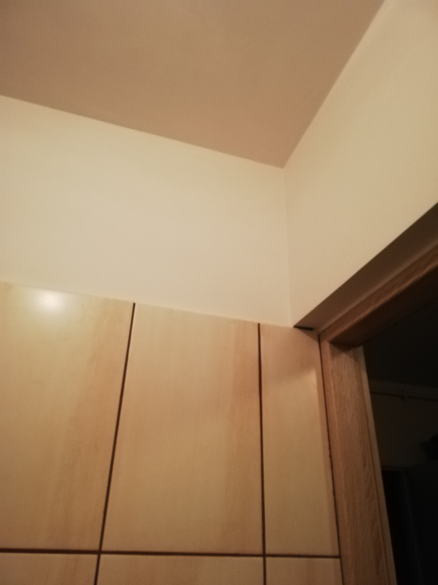 Beżowe płytki na ścianie w rogu pomieszczenia, stykające się z kremową ścianą i drewnianą futryną drzwiową, widok z dołu.