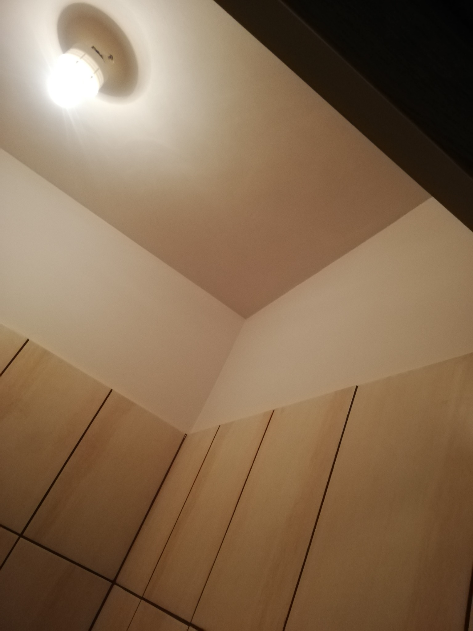 Pomieszczenie po malowaniu: kremowe ściany, fragment sufitu w odcieniu pudrowego różu, widoczna lampa sufitowa i fragment zabudowy meblowej w jasnym drewnie.