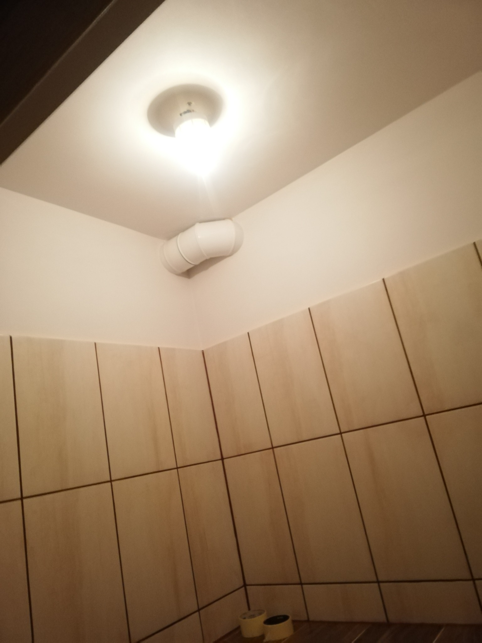 Wykończona łazienka z beżowymi płytkami kwadratowymi, widoczny fragment sufitu z lampą i wentylacją.